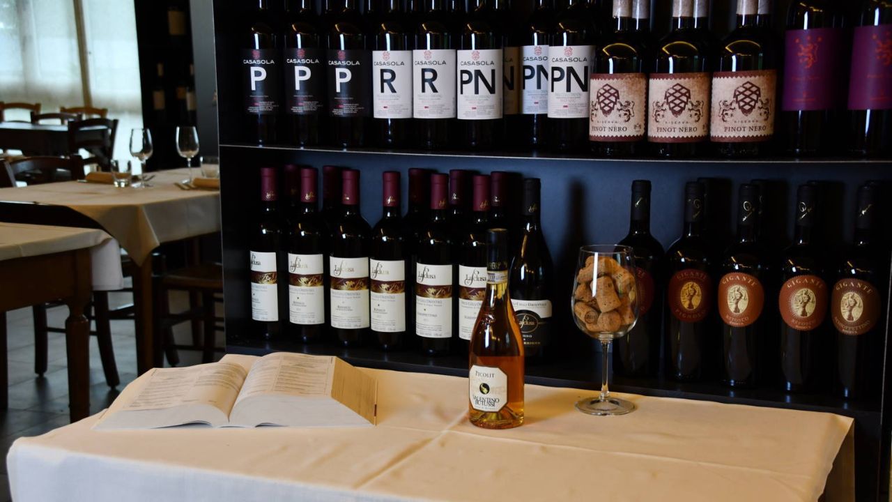 Raffinata selezione di vini esposti con cura all’Osteria Solder. Bottiglie pregiate, un calice colmo di tappi e un libro aperto raccontano la passione per l’enologia e l’arte dell’ospitalità autentica.
